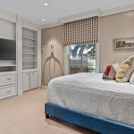 A cozy bedroom with a neatly made bed, blue upholstered frame, multiple pillows, a wooden nightstand, built-in shelves, a flat-screen TV, and a window with brown checkered blinds letting in natural light.