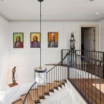 A bright stairway with a black railing, curved wooden steps, large arched window, white paneled walls, three colorful portraits, a glass pendant light, and two sculptures decorating the space.