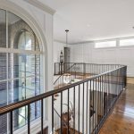 A bright hallway with dark wooden floors, white paneled walls, large windows, and black metal railing overlooking a lower level and a brick wall with arched windows.