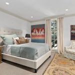 A cozy bedroom with a beige upholstered bed, light blue bedding, wooden nightstands with black lamps, a patterned rug, an armchair, large windows with curtains, and two colorful paintings on the walls.