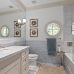 A bright bathroom with marble countertops and walls, a large mirror, two round windows, framed flower prints, a toilet, towel rack, and a bathtub with a beige and gray patterned rug on the floor.