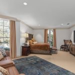 A spacious bedroom with a brown leather sofa, patterned pillows, a blue rug, a large bed with gold bedding, wooden furniture, and a wall-mounted flat-screen TV. Large windows let in natural light.