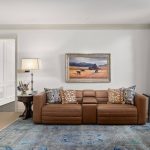 A living room with a brown leather sofa, patterned pillows, a blue rug, a floor lamp with a light shade, a framed landscape painting on the wall, and tall windows with gold curtains letting in natural light.