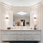 Elegant bathroom with striped wallpaper, white cabinets, double sinks, a large mirror, and two wall-mounted lights. Decorative items, including a silver sculpture and a tissue box, sit on the marble countertop.