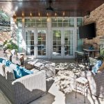Outdoor patio with wicker furniture, blue and neutral cushions, a wall-mounted TV, potted plants, and string lights under a covered roof. Brick walls and glass doors lead inside. Shadows from trees cover the tiled floor.