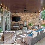 Covered patio with wicker and metal furniture, a TV mounted on a brick wall, potted plants, a ceiling fan, and a small water feature, overlooking a green lawn with trees and wooden benches.