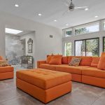 A bright living room with large windows, orange sectional sofas, an orange ottoman, patterned pillows, gray tiled floor, and outdoor greenery visible through glass doors.