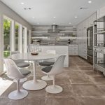 Modern kitchen with gray cabinetry, round white dining table, four white chairs, large tile flooring, open shelving units, and sliding glass doors opening to a backyard with a pool and green lawn.