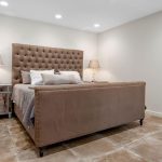 A modern bedroom with a large brown upholstered bed, two bedside tables with matching lamps, neutral walls, recessed ceiling lights, and tiled flooring. The room appears clean and minimalist.