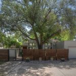 A modern house with a flat roof, large windows, and a white exterior is partly hidden behind a wooden fence and gate, with a large leafy tree in the front yard and a two-car garage on the right side.