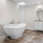 Modern bathroom with a freestanding white bathtub, large tiled floor, double sink vanity with white cabinets, round mirrors, and a glass pendant light. Neutral color palette and clean, minimalist design.