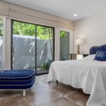 A modern bedroom with large windows, a sliding glass door, a blue velvet chaise lounge, a matching upholstered bed, white bedding, a nightstand with a lamp, and tiled floors. Greenery is visible outside.
