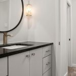 Modern bathroom with white cabinets, black countertop, under-mount sink, gold faucet, round mirror, and wall-mounted light fixture. A hallway with tiled floor is visible through an open doorway.