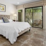 A modern bedroom with a beige upholstered bed, white bedding, bedside tables with lamps, tiled floor, and large sliding glass doors opening to a small outdoor area with greenery and a white wall.