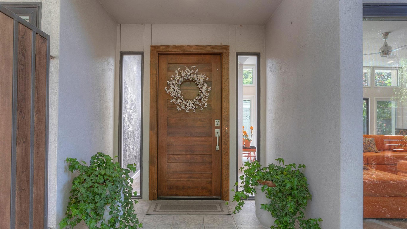 A wooden front door with a white floral wreath, flanked by two tall narrow windows, two potted green plants on either side, and a glimpse of a living room with an orange sofa inside.