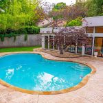 A curved swimming pool with clear blue water is surrounded by a stone patio and landscaped greenery, next to a modern house with large windows and a tree with red leaves by the pool.