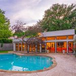 Modern house with large windows and bright interior lights, facing a curvy swimming pool surrounded by stone tiles. Trees and greenery are in the background, and potted plants decorate the patio area. Evening sky overhead.