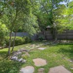 A backyard with lush green grass, several tall trees, and a wooden fence. Stone pavers form a winding path through the yard, and sunlight filters through the leafy branches, creating a peaceful, shaded atmosphere.