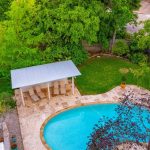 Aerial view of a backyard with a curvy swimming pool, stone patio, three lounge chairs under a covered area, green lawn, potted plant, and surrounded by lush trees.