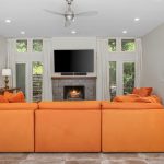 A modern living room with a bright orange sectional sofa facing a fireplace and wall-mounted TV, flanked by tall windows. There are lamps and decor on a console table, and a wooden door is visible on the right.