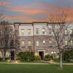 A three-story brick townhouse with lit windows, surrounded by leafless trees and green lawns, is shown at sunset with a colorful sky in the background.