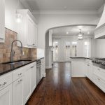 Modern kitchen with white cabinets, dark countertops, tile backsplash, stainless steel sink, gas stove, and wood flooring. An arched doorway leads to a bright dining area with large windows and glass doors.
