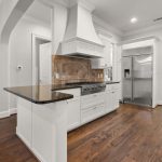 Spacious modern kitchen with white cabinets, stainless steel appliances, a large refrigerator, dark granite countertops, tile backsplash, hardwood floors, and arched doorways leading to adjacent rooms.
