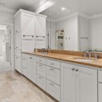 A modern bathroom with white cabinets, a brown marble countertop, a large mirror, and beige tile floors. There is a single sink, chrome fixtures, and a walk-in closet visible through an open doorway.