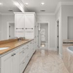 Spacious bathroom with a long marble countertop, double sinks, large mirrors, white cabinets, a built-in bathtub with tile accents, and beige tiled floors. The space is bright and modern with neutral colors.