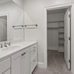 White bathroom with a single sink vanity, large mirror, and silver fixtures. An open door leads to a walk-in closet with built-in shelves and hanging rods. The space has clean, modern lines and neutral colors.