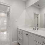 A modern bathroom with white cabinets, a double sink vanity, a large mirror, and silver fixtures. An open doorway leads to a bright hallway with arched doorways and light-colored floors.