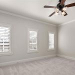Empty bedroom with light gray walls, three shuttered windows, white trim, double closet doors, and a ceiling fan with lights. The room is carpeted and lit with natural light from the windows.