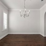 A bright, empty room with white walls, dark wooden floors, a large window with white shutters, a decorative chandelier, and an arched doorway leading to another area.