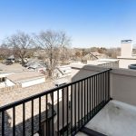 View from a balcony with a black metal railing, overlooking rooftops, garages, leafless trees, and a clear blue sky in a suburban neighborhood.