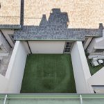 A view from above of a small, enclosed courtyard with artificial grass, surrounded by white walls. Patio furniture is visible in two corners, and rooftops cast shadows over the area.