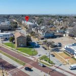 Aerial view of a suburban neighborhood with houses, a church, businesses, and cars on the street; a red location pin marks a specific building in the background.