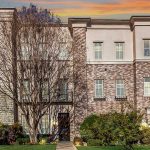 A row of modern townhouses with brick and stone facades, large windows, and trimmed landscaping, set against a colorful sunset sky. A leafless tree stands in front of one townhouse.