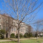 A row of tan townhouses lines a suburban street with leafless trees and neatly trimmed lawns. Parked cars are visible along the curb under a clear blue sky.
