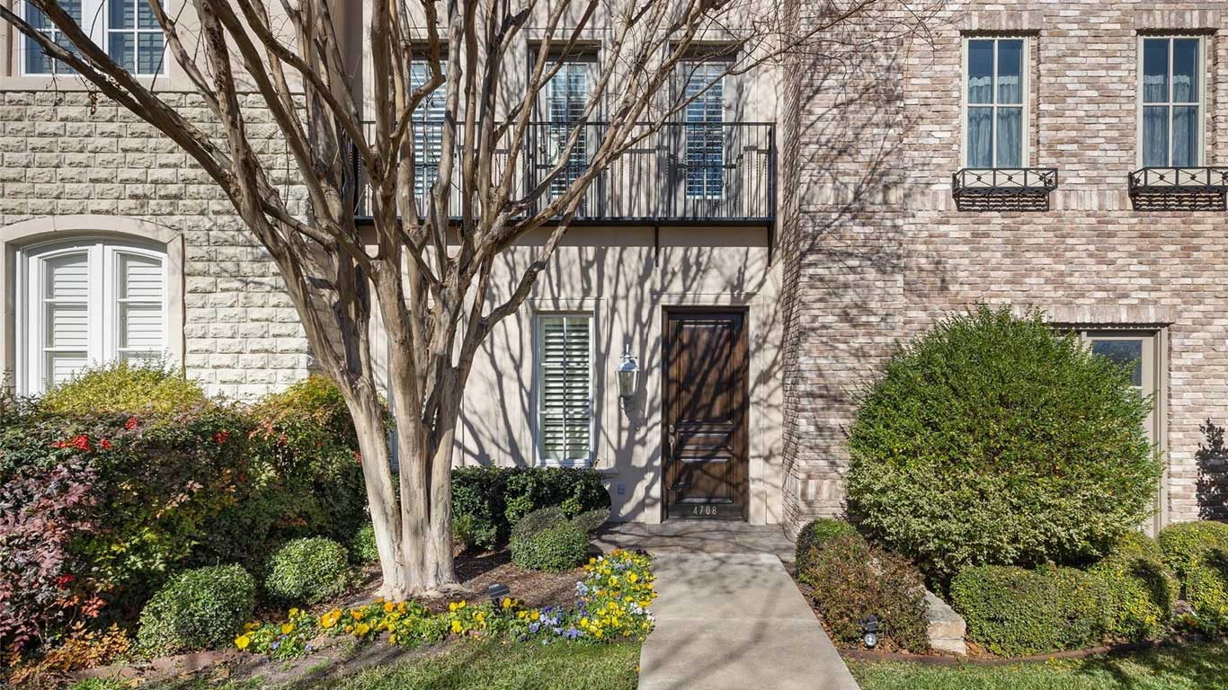 A townhouse entrance with a dark wooden door, large front window with shutters, leafless tree, trimmed bushes, and colorful flowers along a paved walkway against a stone and brick façade.