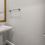 A minimalist bathroom with a white pedestal sink, a silver faucet, a gold-framed mirror above the sink, a chrome towel bar on the wall, and a white door slightly open. The walls are light gray.