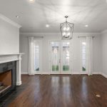 A bright, empty living room with hardwood floors, a white fireplace on the left, double French doors with sheer curtains leading outside, and a modern geometric light fixture on the ceiling.