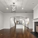 A spacious, modern living room with dark wood floors, white walls, a black-tiled fireplace, a large lantern-style ceiling light, and an open view into a bright kitchen with an island and white cabinetry.