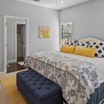 A cozy bedroom with a large bed featuring blue and white floral bedding, yellow pillows, and a navy tufted bench at the foot. There are side tables, lamps, a cushioned chair, and wall art, with sunlight streaming through the windows.