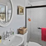 Bright bathroom with white tile walls, a glass shower door, a round mirror above a sink, pink towels, a potted cactus, and a window with horizontal blinds letting in natural light.