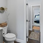 A bright bathroom with a gray vanity, sink, toilet, basket, and a round wall mirror. An open door reveals a hallway and a bedroom with a bed and colorful rug in the background.