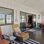 Covered patio with cushioned chairs, floral pillows, a small side table, and a view into a living room through large sliding glass doors. Windows overlook a grassy yard with a white fence.
