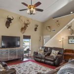 A spacious living room with brown leather sofas, a large TV, and multiple mounted animal heads on beige walls. A ceiling fan hangs from a high ceiling, with stairs leading to an upper level and windows letting in natural light.