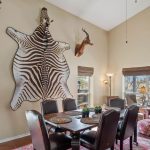 A dining area with a table and six chairs, surrounded by brown armchairs. Large zebra hide and mounted antelope heads decorate the beige walls. Sunlight streams through windows showing a natural, grassy landscape outside.
