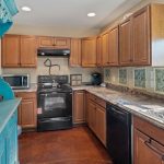 A kitchen with wooden cabinets, black appliances, marble-patterned countertops, a double sink by a window, and a turquoise hutch with glassware and bottles on the left side.