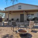 A metal house with a covered porch, outdoor chairs, ceiling fans, and a fire pit in the yard, surrounded by trees and a fence under a clear blue sky.